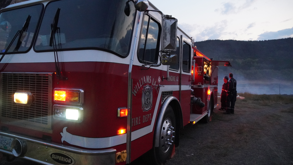 A fire truck supports a wildfire, with a large red engine siting to the right, and a few firefighters stand behind it to the right with smoke burning in a field in the foreground. Thankfully this truck is the proper colour.